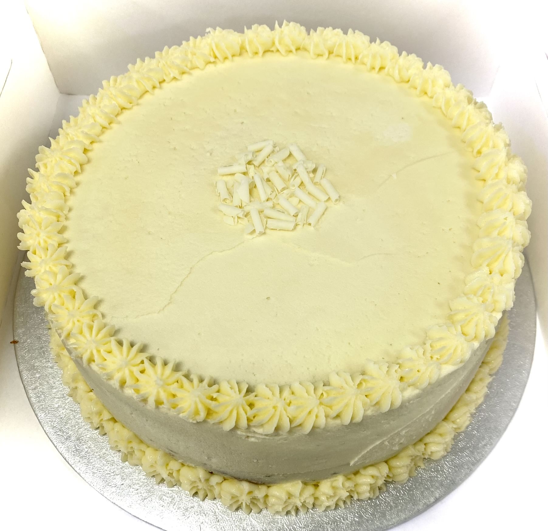 A round Vanilla Gateaux with white frosting is displayed inside a white box. This classic cake is adorned with small piped rosettes along the top edge and features a cluster of white chocolate shavings in the center. A small crack is visible on the surface of this exquisite dessert's frosting.